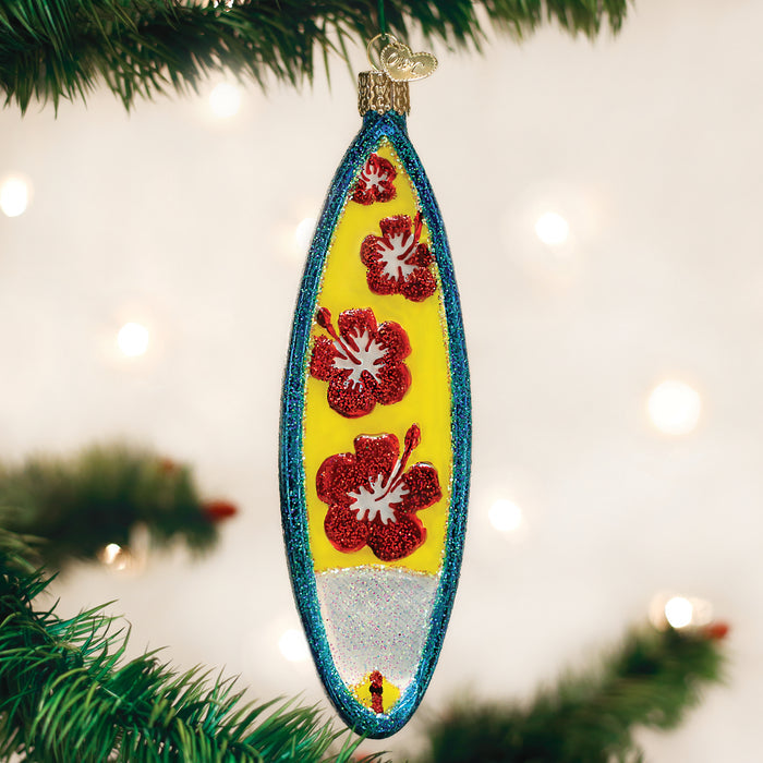 A glittery surfboard-shaped Christmas ornament decorated with red hibiscus flowers hangs on a green pine tree branch, with blurred white lights in the background.